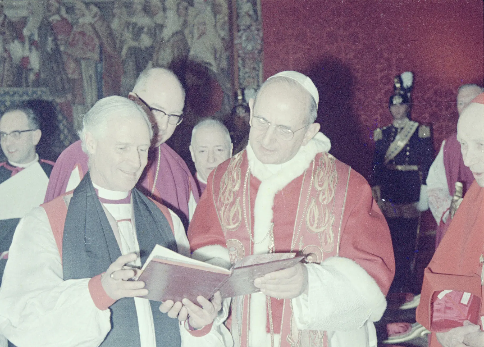 Bishop John Moorman, leader of the Anglican delegation at the Second Vatican Council, examines a book with Pope Paul VI. Archbishop Jan Willebrands peers over Moorman's shoulder, while Cardinal Augustin Bea watches from the pope's side